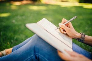 Image of someone from the waist down sitting on grass with a book on their lap and writing.